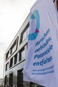 MV-Ostseekooperation MV-Ostseestrategie Fahne Eine weiße Fahne mit dem Logo und Slogan der „MV-Ostseestrategie“ („Menschen verbinden. Potentiale entfalten.“) weht im Vordergrund vor der modernen Außenfassade des Veranstaltungsgebäudes.