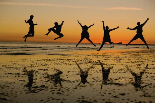 Jugendliche springen am Strand bei Sonnenuntergang Silhouetten von fünf jungen Menschen, die bei Sonnenuntergang am Strand in die Luft springen, mit Spiegelungen im nassen Sand.